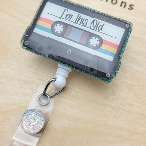 May include: A green glitter cassette tape with a rainbow stripe design and the text "I'm This Old" on a white background. The cassette tape is attached to a white retractable badge holder with a clear plastic strap and a sparkly iridescent button.