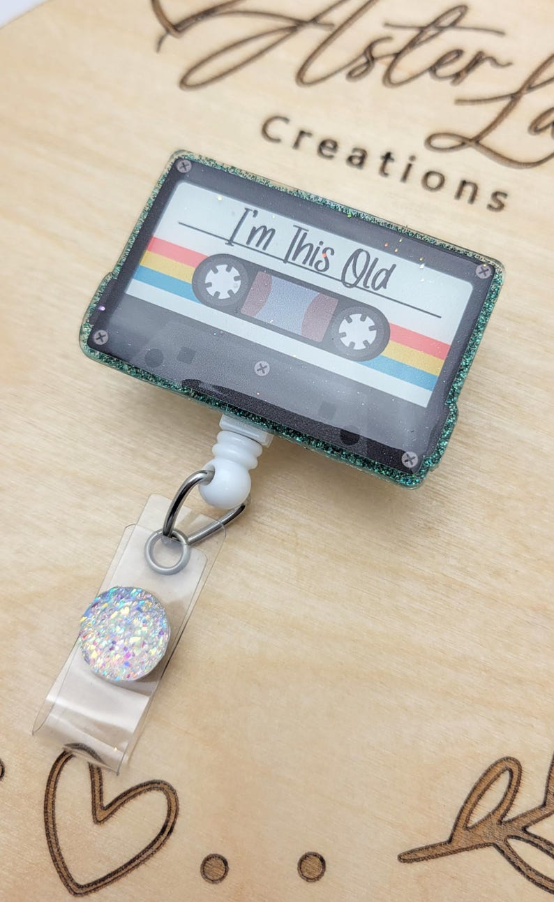 May include: A green glitter cassette tape with a rainbow stripe design and the text "I'm This Old" on a white background. The cassette tape is attached to a white retractable badge holder with a clear plastic strap and a sparkly iridescent button.