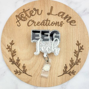 May include: A white and black glitter badge reel with the text "EEG Tech" on it. The badge reel is attached to a wooden circle with the text "Aster Lane Creations" engraved on it.