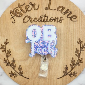 May include: A white glitter badge holder with the text "OB Tech" in white letters on a purple glitter background. The badge holder is attached to a clear plastic retractable badge reel with a silver glitter circle.