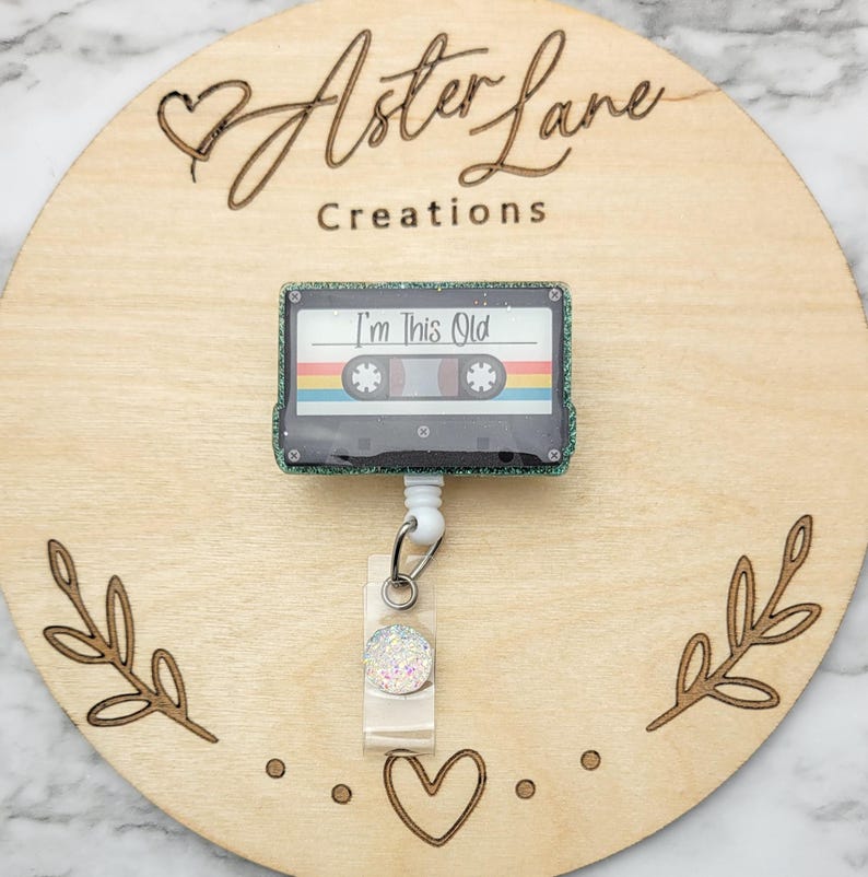 May include: A green glitter cassette tape with the text "I'm This Old" on it. The cassette tape is attached to a white retractable badge reel with a clear plastic cover and a sparkly white circle.