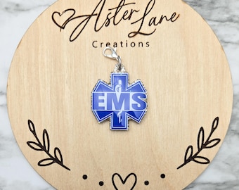 May include: A blue and silver EMS charm with the text "EMS" on a wooden round with the text "Aster Lane Creations". The charm is attached to a silver clasp. The wooden round has a heart and leaf design.