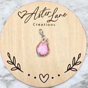 May include: A pink charm with the text "Hold on, let me overthink this" in a flame shape, adorned with flowers and rhinestones. The charm is attached to a silver clasp. The charm is displayed on a wooden round board with the text "Aster Lane Creations".