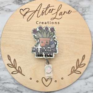 May include: A glittery badge reel featuring a cartoon succulent in a terracotta pot holding a watering can. The text reads I'M ROOTING FOR YOU in bold black letters. The design is mounted on a clear plastic clip with a small circular glitter accent.