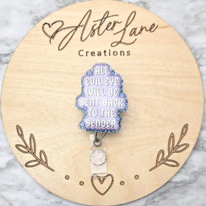 May include: A badge reel with the text "All Evil Eye Will Be Sent Back To The Sender" in a light purple color with glitter accents. The badge reel is on a wooden background with the words "Aster Lane Creations" and decorative leaf and heart designs.