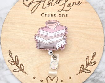 May include: A badge reel featuring a stack of books with a coffee cup on top. The badge reel has a glittery border and a clear plastic clip. The wooden background has the text "Aster Lane Creations".