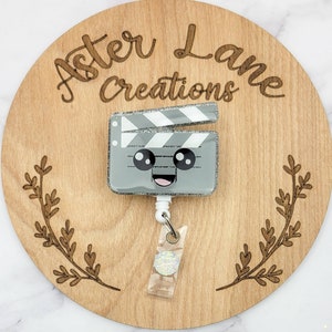 May include: A gray and white glitter film clapboard with a cute cartoon face. The clapboard is attached to a retractable badge reel with a clear plastic badge holder and a silver glitter circle.