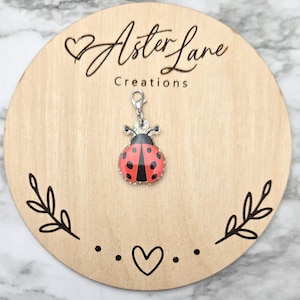 May include: A ladybug charm with red and black enamel, accented with clear crystals, hangs from a silver-tone clasp. The charm is displayed on a round wooden plaque with the text "Aster Lane Creations". The plaque has a decorative leaf and heart design.