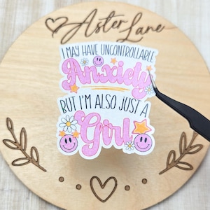 May include: A round wooden sign with the text "Aster Lane" in cursive script. A sticker with the words "I MAY HAVE UNCONTROLLABLE Anxiety BUT I'M ALSO JUST A Girl" is centered on the sign. The sticker has a pink and white color scheme with smiley faces and stars.