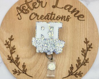 May include: A white glitter badge reel with the letters "RT" and the text "Respiratory Therapist". The badge reel is on a wooden background with the text "Aster Lane Creations".