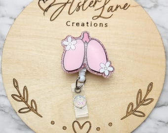 May include: Pink glitter resin lungs with white flowers and a white glitter retractable badge reel. The reel has a clear plastic badge holder and a white glitter charm. The background is a light brown wooden circle with the text "Aster Lane Creations" in black.