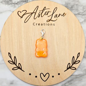 May include: An orange gummy bear charm with a silver clasp hangs on a wooden round. The charm has a glittery outline and a translucent appearance. The wooden round has the text "Aster Lane Creations" and decorative leaf and heart designs.