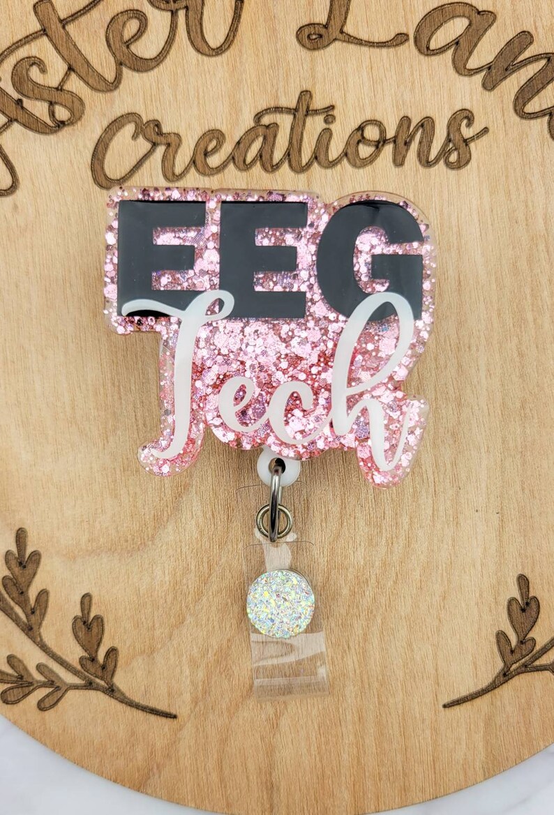 May include: Pink glitter badge reel with the text "EEG Tech" in black and white lettering. The reel has a clear plastic backing and a white glitter charm.