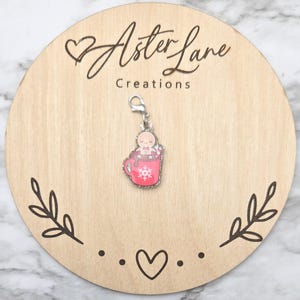 May include: A charm featuring a gingerbread man in a pink mug with a snowflake design. The charm is attached to a silver clasp and is displayed on a wooden round with the text "Aster Lane Creations".