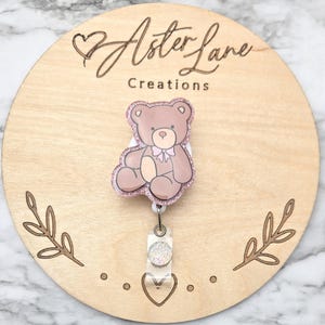 May include: A brown teddy bear badge reel with a pink glitter outline and bow. The badge reel is attached to a clear plastic holder with a glittery accent. The wooden background has the text "Aster Lane Creations".