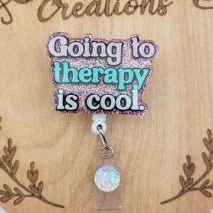 Op de afbeelding: Een roze, turquoise en gouden glitter badgehouder met de tekst "Going to therapy is cool." De badgehouder is bevestigd aan een witte intrekbare badgehaspel met een doorzichtige plastic badgehouder met een glinsterende regenboogkleurige cirkel.