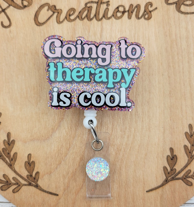 Op de afbeelding: Een roze, turquoise en gouden glitter badgehouder met de tekst "Going to therapy is cool." De badgehouder is bevestigd aan een witte intrekbare badgehaspel met een doorzichtige plastic badgehouder met een glinsterende regenboogkleurige cirkel.