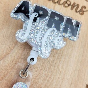 May include: A silver glitter badge reel with a black and white "APRN Life" design. The reel has a clear plastic case and a silver glitter charm.