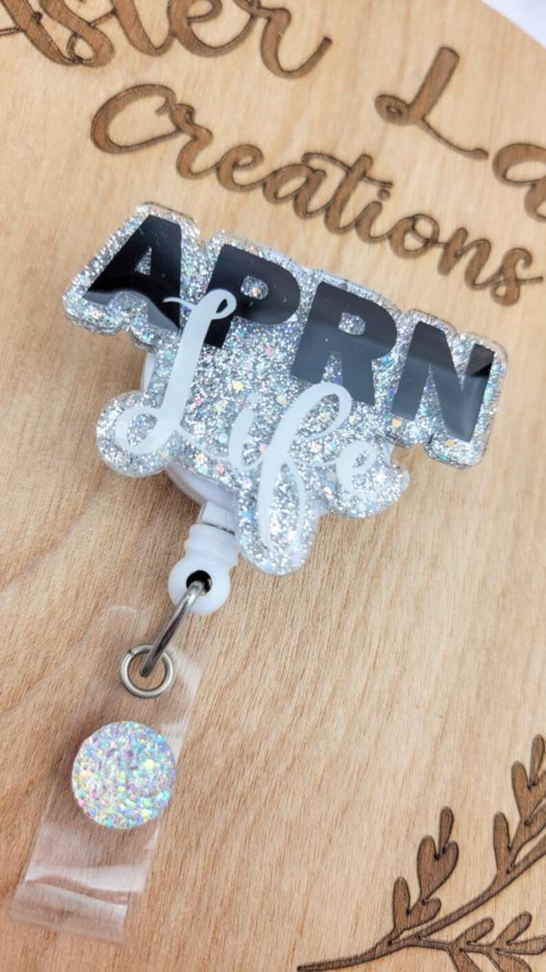 May include: A silver glitter badge reel with a black and white "APRN Life" design. The reel has a clear plastic case and a silver glitter charm.