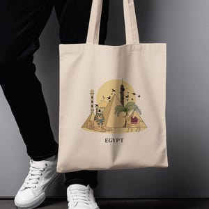 May include: Beige tote bag featuring an illustration of Egyptian landmarks, including pyramids, a sphinx, and a camel. The design is set against a yellow sun with flying birds, and the word "EGYPT" is printed below.