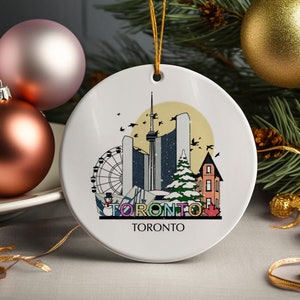 Könnte beinhalten: Runder weißer Keramik-Weihnachtsschmuck mit einem Toronto-Skyline-Design. Das Ornament zeigt den CN Tower, ein Riesenrad und einen Schneemann. Das Wort "TORONTO" ist in bunten Buchstaben geschrieben. Ein goldener Faden zum Aufhängen ist befestigt.