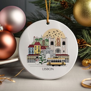 May include: White ceramic Christmas ornament featuring a colorful illustration of Lisbon, Portugal, with a yellow tram, buildings, and the text "LISBON". The ornament has a gold string for hanging and is surrounded by Christmas decorations.