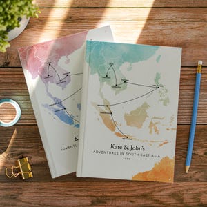 May include: Two hardback journals featuring a watercolour map of South East Asia with black lines connecting locations including Laos, Thailand, Vietnam, Cambodia, Malaysia, Singapore, Indonesia, and Philippines. Text reads Kate & John's Adventures in South East Asia 2026.