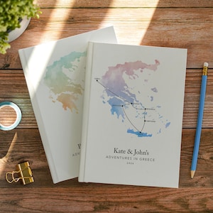 Könnte beinhalten: Zwei weiße Hardcover-Notizbücher mit einer Aquarellkarte von Griechenland. Das obere Notizbuch zeigt eine schwarze Linie, die Orte wie Korfu, Nafplio, Athen, Mykonos, Santorin und Kreta verbindet, mit dem Text Kate & John's ADVENTURES IN GREECE 2026.
