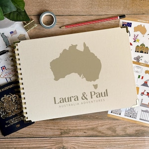 May include: A cream-colored travel journal with a gold spiral binding and the outline of Australia. The journal is personalized with the names "Laura & Paul" and the text "Australia Adventures". Other travel-themed items are in the background.