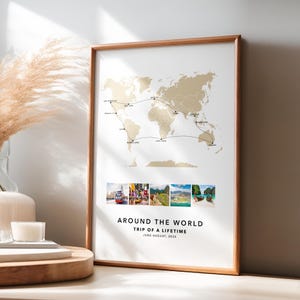 May include: A framed world map print with a travel route marked in black. The map is in gold tones, with a series of small photos below. Text reads "AROUND THE WORLD TRIP OF A LIFETIME JUNE-AUGUST, 2023". The frame is a light brown color.