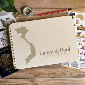 May include: A cream-coloured travel journal with a gold spiral binding. The cover features a map of Vietnam and the names "Laura & Paul" with the text "Vietnam Adventures". A passport and travel stickers are visible.