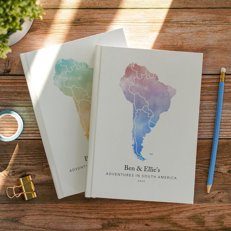 May include: Two white hardcover journals with watercolor map designs of South America. One journal has a rainbow-colored map, the other a red, purple, and blue map. Text on the journals reads "Ben & Ellie's Adventures in South America 2025" and "Adventi". A blue pencil rests nearby.