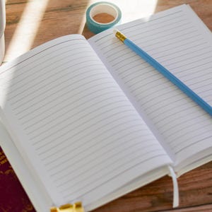 May include: An open, lined notebook with a blue pencil and a roll of teal washi tape on a wooden surface. The notebook's pages are white with horizontal lines. The pencil has a yellow eraser. The tape is teal with a white pattern.