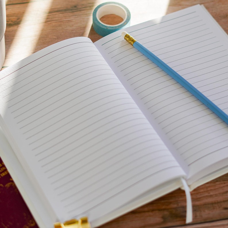 May include: An open, lined notebook with a blue pencil and a roll of teal washi tape on a wooden surface. The notebook's pages are white with horizontal lines. The pencil has a yellow eraser. The tape is teal with a white pattern.
