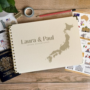 Può includere: Un diario di viaggio color crema con rilegatura a spirale dorata, con il testo "Laura & Paul Japan Adventures" e una mappa del Giappone. Sono visibili anche un passaporto, adesivi e una matita.