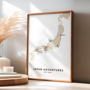 May include: A framed print titled "Japan Adventures" with a map of Japan in beige tones. The map highlights a travel route with city names like Tokyo, Kyoto, and Hiroshima. The frame is a light brown color, and the print includes the date "May 2024".