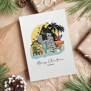 May include: A Christmas card with a Hawaiian theme. The card features a colorful illustration of a beach scene with palm trees, buildings, and the word "Aloha." The text "Merry Christmas Hawaii" is written below the illustration.