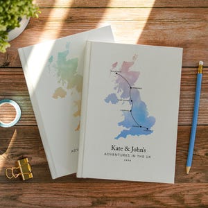 Könnte beinhalten: Zwei weiße Hardcover-Notizbücher mit einer Aquarellkarte des Vereinigten Königreichs. Das vordere Notizbuch zeigt eine schwarze Linie, die Isle of Skye, Edinburgh, Lake District, Liverpool, Oxford und London verbindet, mit dem Text Kate & John's ADVENTURES IN THE UK 2026.