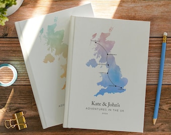 May include: Two white hardcover journals featuring a watercolor map of the UK. The front journal displays a black line connecting Isle of Skye, Edinburgh, Lake District, Liverpool, Oxford, and London, with the text Kate & John's ADVENTURES IN THE UK 2026.