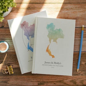 May include: Two white hardcover journals featuring a watercolor map of Thailand. The top journal displays a multicolored map with the text James & Molly's ADVENTURES IN THAILAND 2025. The bottom journal shows a map with a black travel route line.