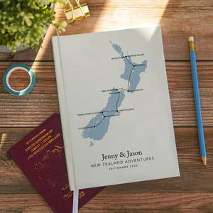 May include: A white journal with a map of New Zealand and travel route details in blue. The journal is personalized with the names "Jenny & Jason" and the text "New Zealand Adventures, September 2023". A blue pencil and passport are nearby.
