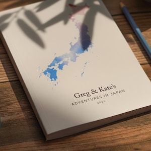May include: A white journal with a watercolor map of Japan in blue and purple hues. The cover reads "Greg & Kate's Adventures in Japan 2025". A blue pencil rests on a wooden surface next to the journal.