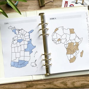 May include: Open planner with pages featuring hand-drawn maps of the USA and Africa. The USA map shows states colored in blue, while the African map highlights countries in brown. The planner is open to display the maps.