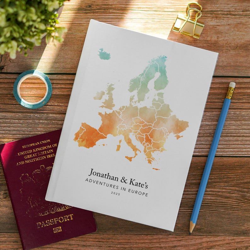 May include: A white journal with a watercolor map of Europe in orange, green, and blue. The journal is personalized with the text "Jonathan & Kate's Adventures in Europe 2025." A blue pencil, gold binder clip, and passport are also visible.