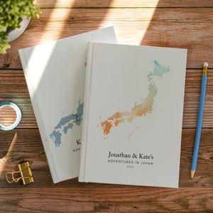 May include: Two white hardcover journals featuring a watercolor map of Japan. The top journal displays a gradient map and the text "Jonathan & Kate's ADVENTURES IN JAPAN 2025". The bottom journal shows a blue map with "KYOTO" and "OSAKA" labels.
