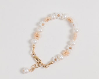 May include: A delicate bracelet made with white and peach colored flower beads and pearl accents. The bracelet has a gold clasp.