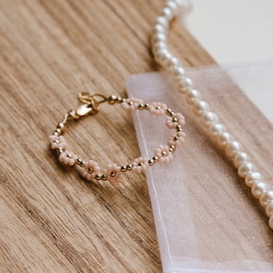 May include: A delicate gold bracelet with pink flower beads. The bracelet is lying on a white fabric surface with a pearl necklace.