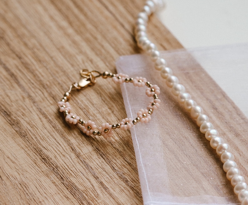 May include: A delicate gold bracelet with pink flower beads. The bracelet is lying on a white fabric surface with a pearl necklace.