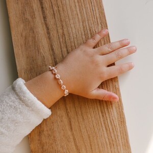 May include: A child's wrist wearing a delicate bracelet with small pink flower charms. The bracelet is made of gold chain and the flowers are made of beads.