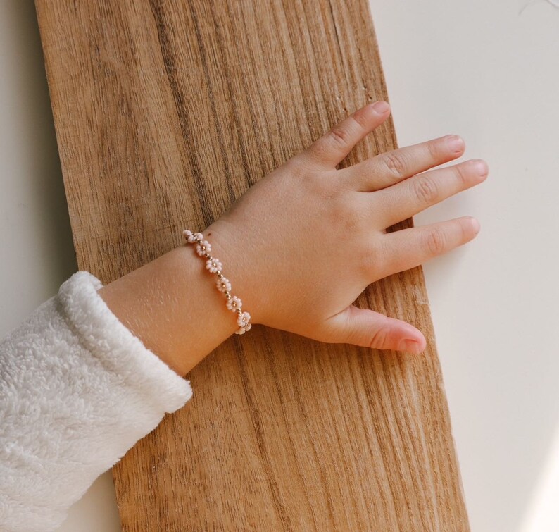 May include: A child's wrist wearing a delicate bracelet with small pink flower charms. The bracelet is made of gold chain and the flowers are made of beads.